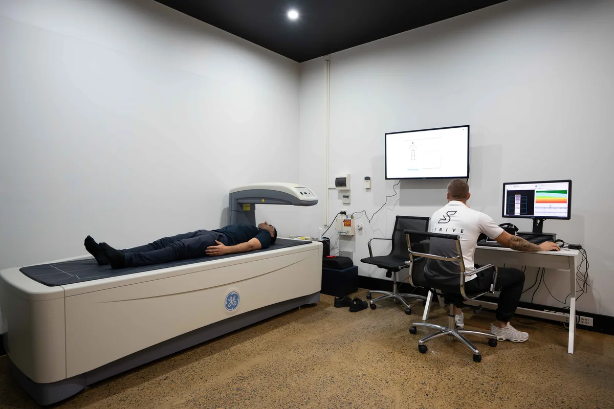 A person lying on a DEXA scanning table while an operator works at the computer at STRIVE.